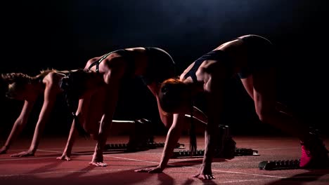 Tres-chicas-en-ropa-de-color-negro-están-en-las-almohadillas-de-partida-para-iniciar-la-carrera-en-la-competencia-a-la-luz-de-las-luces-y-correr-hacia-la-meta