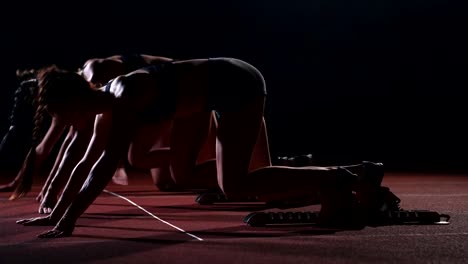 Tres-chicas-en-ropa-de-color-negro-están-en-las-almohadillas-de-partida-para-iniciar-la-carrera-en-la-competencia-a-la-luz-de-las-luces-y-correr-hacia-la-meta