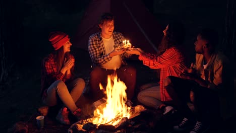 Excited-tourists-are-congratulating-young-man-on-birthday-giving-him-cake,-guy-is-making-wish-with-closed-eyes-and-blowing-candles,-his-friends-are-clapping-hands.