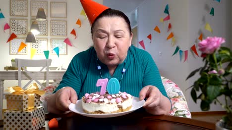 Mujer-senior-feliz-sosteniendo-pastel-a-cámara-celebrando-cumpleaños-con-su-familia-a-través-de-video-chat