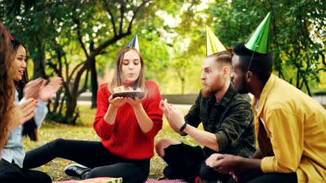 Hermosa-chica-en-sombrero-del-partido-celebra-cumpleaños-con-amigos-en-Parque-deseo-hacer-picnic,-soplar-velas-sobre-el-pastel-y-divertirse-riéndose.-Concepto-naturaleza-y-eventos.