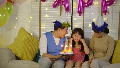 A-cute-little-asian-girl-celebrating-her-birthday-with-a-cake-and-happy-family.-She-gives-a-big-smile-and-her-family-applaud-her.-In-slow-motion.