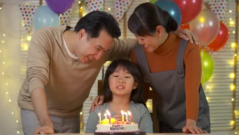 A-cute-little-asian-girl-sitting-at-table-and-blowing-candles-on-birthday-cake-while-her-family-standing-behind-and-sing-a-song-to-her.-She-gives-a-big-smile-and-her-family-applaud-her.-In-slow-motion.