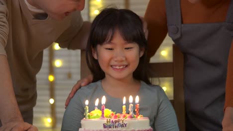 A-cute-little-asian-girl-sitting-at-table-and-blowing-candles-on-birthday-cake-while-her-family-standing-behind-and-sing-a-song-to-her.-She-gives-a-big-smile-and-her-family-applaud-her.-In-slow-motion.