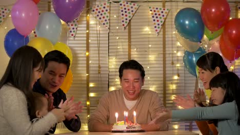 Happy-asian-male-sitting-at-table-surrounded-by-his-family-and-friends-celebrates-his-birthday-with-a-cake.