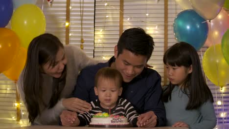 A-cute-little-boy-sitting-at-table-and-blowing-candles-on-birthday-cake-while-his-family-standing-behind-and-sing-a-song-to-him.-In-slow-motion.