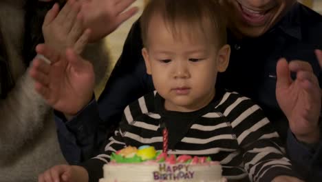 A-cute-little-boy-sitting-at-table-and-blowing-candles-on-birthday-cake-while-his-family-standing-behind-and-sing-a-song-to-him.-In-slow-motion.