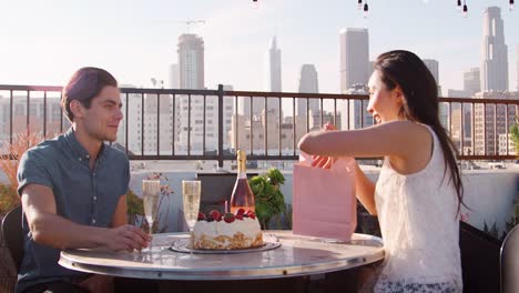 Mann-Frau-Geschenk-und-Karte-zu-geben,-wie-sie-auf-Dachterrasse-mit-Skyline-der-Stadt-im-Hintergrund-feiern