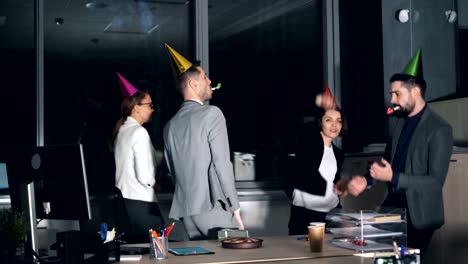 Oficinistas-de-felices-jóvenes-bailan-en-la-fiesta-de-empresa-con-sombreros-de-fiesta-y-soplando-cuernos.-Caja-de-regalo-y-torta-de-cumpleaños-son-visibles-en-el-escritorio.