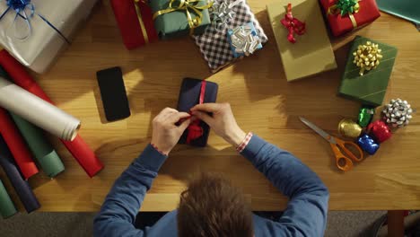 Top-View-of-Young-Man-Skillfully-Packing-Colorful-Gifts-in-His-Sunny-Studio.-Smartphone-with-Green-Mock-Up-Screen-Lies-on-the-Table.