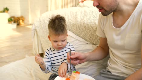 Pequeño-niño-adorable-celebrando-su-cumpleaños-con-el-joven-padre-comer-pastel-en-el-dormitorio