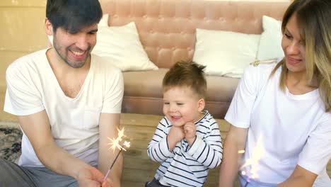 Young-father-and-mother-celebrate-their-son-birthday-burning-sparklers-at-home-and-smiling