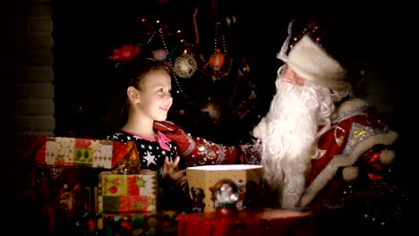 In-der-Dämmerung-der-Nacht,-eine-hübsche-blonde-Mädchen-mit-rosa-Schleife-im-Haar,-in-einem-ein-schönes-Kleid-bewundert-Weihnachtsgeschenke,-öffnet-sich-eine-große-Kiste,-erfüllt-den-Raum-mit-einem-magischen-Glanz,-sie-ist-glücklich,-froh.-Weihnachtsmann-sitzt-in-ihrer-Nähe.-In-der-backgr