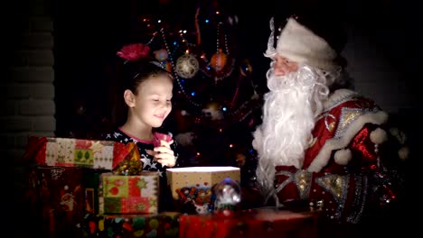 In-der-Dämmerung-der-Nacht,-eine-hübsche-blonde-Mädchen-mit-rosa-Schleife-im-Haar,-in-einem-ein-schönes-Kleid-bewundert-Weihnachtsgeschenke,-öffnet-sich-eine-große-Kiste,-erfüllt-den-Raum-mit-einem-magischen-Glanz,-sie-ist-glücklich,-froh.-Weihnachtsmann-sitzt-in-ihrer-Nähe.-In-der-backgr