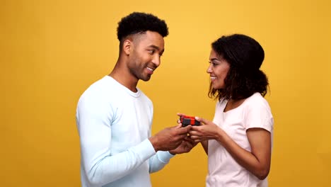 Smiling-young-african-man-giving-a-present-box-to-his-girlfriend-isolated-over-yellow-background