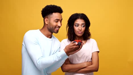 Smiling-young-african-man-giving-a-present-box-to-his-upset-grumpy-girlfriend-isolated-over-yellow-background