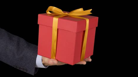 Mans-hand-in-a-business-suit-holds-a-red-gift-box-with-a-gold-bow.-Hand-with-a-gift-appears-in-the-frame.-Red-gift-box.-A-businessman-gives-a-gift.-Alpha-channel-chroma-key-transparent-background.