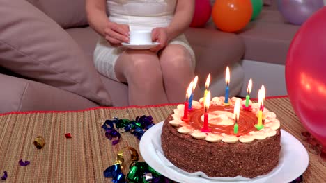 Woman-with-cup-of-coffee-sitting-on-sofa-near-birthday-party-cake-with-burning-candles