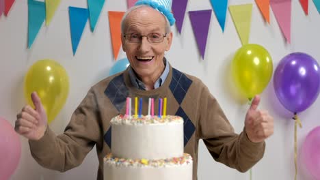 Cheerful-senior-blowing-candles-on-a-birthday-cake-and-holding-his-thumbs-up