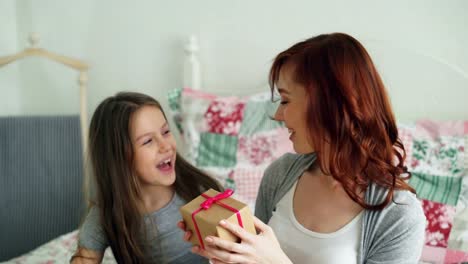 Kleine-süße-Mädchen,-die-ihre-Jungen-glücklich-Mutter-feiern-Muttertag-auf-in-gemütlichen-Schlafzimmer-zu-Hause-sitzen-Geschenkbox-verleiht