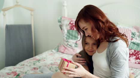 Happy-younf-mother-kissing-her-cute-daughter-presenting-gift-box-on-celebration-sitting-on-bed-in-light-bedroom-at-home