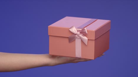 Mano-de-mujer-sosteniendo-una-caja-de-regalo-rosa-en-pantalla-azul;-Concepto-de-celebración