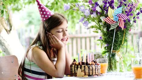 Glückliches-Mädchen-am-Tisch-im-Garten-sitzen-und-bewundert-eine-festliche-Torte