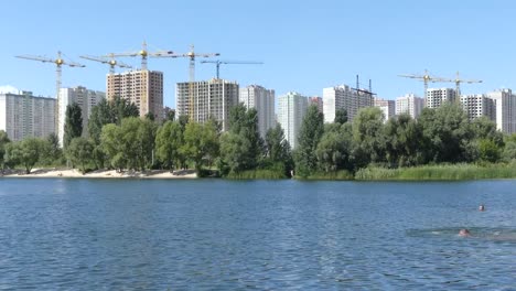 Ein-See-vor-dem-Hintergrund-der-Häuser-im-Bau.-Wolkenkratzer-sind-in-der-Nähe-des-Sees-gebaut.-Bau-von-Wohngebäuden-in-der-Nähe-des-Sees.