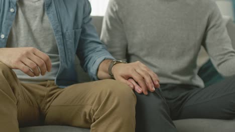 Close-Up-of-a-Gentle-Male-Gay-Couple-Sitting-Together-on-a-Sofa-at-Home.-Boyfriend-Puts-His-Hand-on-Partner's.-They-are-Casually-Dressed-and-Their-Room-Has-Modern-Interior.