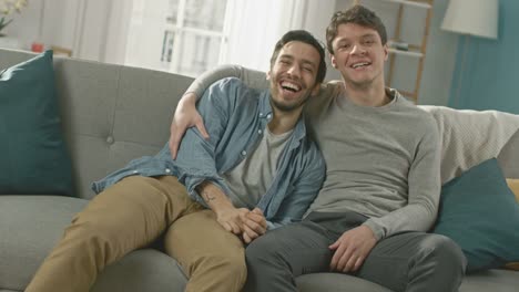 Pareja-Gay-masculino-atractivo-lindo-sentarse-juntos-en-un-sofá-en-casa.-Novios-son-Hugging-y-abrazándose-unos-a-otros.-Son-Joyful-y-ríe.-Son-vestidos-casualmente-y-sala-tiene-un-Interior-moderno.