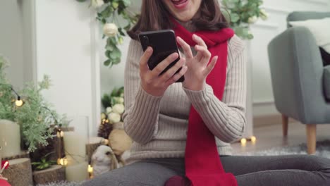 Cheerful-happy-young-Asian-woman-using-smartphone-to-check-social-media-in-her-living-room-at-home-in-Christmas-Festival.-Lifestyle-women-celebrate-Christmas-and-New-year-concept.