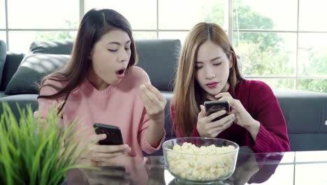 Asiatische-Frauen-mit-Smartphone-und-Essen-Popcorn-im-Wohnzimmer-zu-Hause-Mitbewohner-Freund-Gruppe-Momente-lustige-auf-dem-Sofa-liegend.-Lifestyle-Frauen-entspannen-zu-Hause-Konzept.