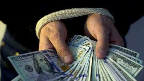 male-hand-bound-with-the-rope-holding-pile-of-money-american-dollar-bank-notes,-financial-rectrictions-concept