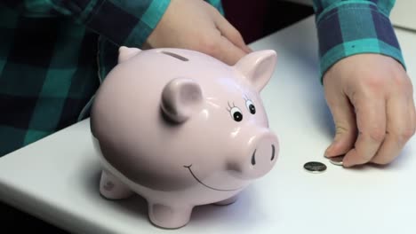 On-the-table-there-is-a-piggy-bank-in-the-form-of-a-pink-pig.-Lying-scattered-coins.-A-man-collects-coins-from-the-table-and-puts-them-in-a-piggy-bank.