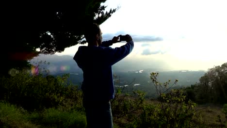 Man-takes-pictures-of-golden-sunset-in-mountains-with-phone-standing-back-close-up-slow-motion.-Photo-memories-of-summer-vacation-with-mobile-camera