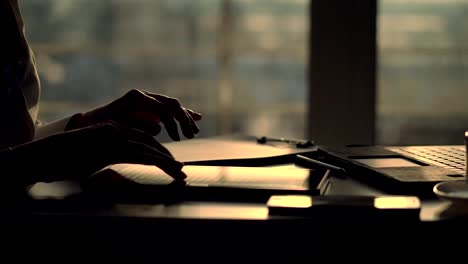 closeup,-dark-silhouette-of-female-hands.-she-is-typing-something-in-tablet,-next-is-a-cup,-mobile,-laptop-on-table.-Blurred-window-background-,-a-ray-of-light-is-reflected-in-the-tablet-screen