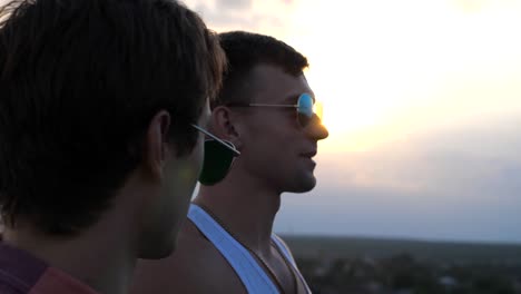 Profile-of-young-male-couple-in-sunglasses-standing-on-the-edge-of-rooftop-and-talking.-Handsome-gay-boys-relaxing-on-roof-of-high-rise-building-and-enjoying-beautiful-cityscape.-Close-up-Slow-motion