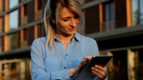 Close-up-portrait-of-a-beautiful-business-woman-holding-and-using-tablet-outside,-girl-looking-at-camera.-Modern-architecture-building-background