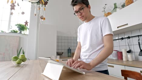 Attractive-man-at-home-using-tablet-in-kitchen-sending-message-on-social-media-smiling-enjoying-modern-lifestyle-wearing-white-shirt