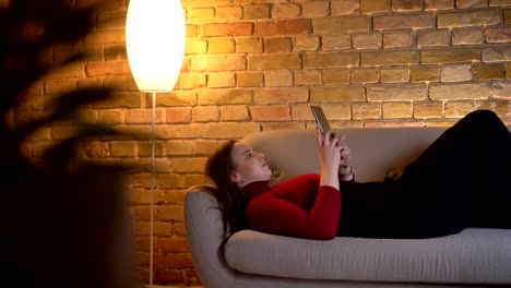 Closeup-shoot-of-young-pretty-caucasian-female-texting-on-the-tablet-while-lying-on-the-couch-indoors