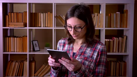 Nahaufnahme-von-jungen-attraktiven-Studentin-in-Brille-mit-dem-Tablet-und-Blick-auf-die-Kamera-lächelnd-in-der-Universitätsbibliothek-drinnen