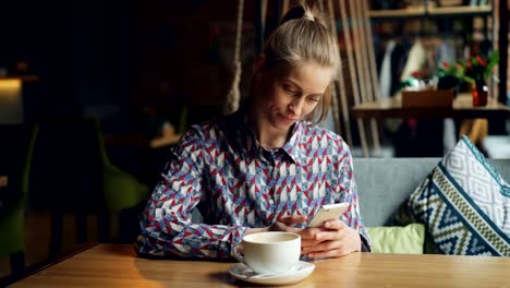 Nettes-Mädchen-mit-Smartphone-in-Café-berühren-Bildschirm-lächelnd-sitzend-am-Tisch