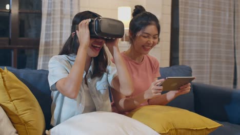 Lesbian-lgbt-women-couple-using-tablet-at-home,-Asian-female-feeling-happy-using-laptop-and-VR-playing-games-together-while-lying-sofa-in-living-room-in-night.-Lover-celebrate-holiday-concept.