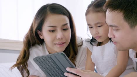 Asiatische-Familie-lacht-und-schaut-in-digitalen-Tablet,-während-auf-dem-Bett-im-Schlafzimmer-liegen