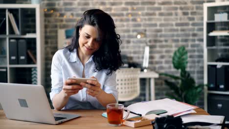 Joyful-girl-playing-video-game-on-smartphone-touching-screen-in-workplace