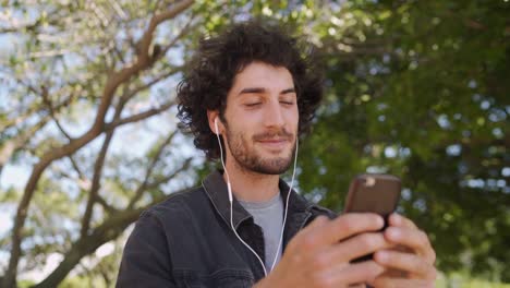 Retrato-de-un-joven-sonriente-con-auriculares-en-los-oídos-charlando-en-las-redes-sociales-en-el-teléfono-inteligente-en-el-parque