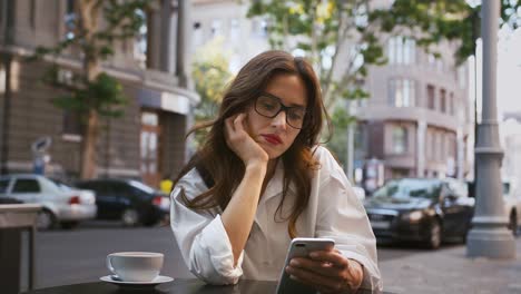 Geschäftsfrau-in-Brille,-weißes-Hemd.-Sitzen-am-Tisch-mit-tasse-Kaffee-im-Straßencafé.-Durchsuchen-von-Nachrichten-auf-dem-Smartphone,-Online-Arbeit.-Nahaufnahme