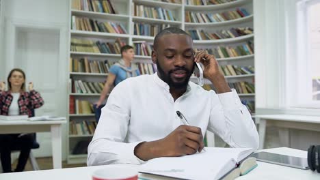 Zeitlupe-von-sympathischen-zufriedenen-bärtigen-afrikanischen-amerikanischen-Kerl,-der-Notizen-in-Copybook-während-seiner-Telefon-Gespräch-in-der-Bibliothek