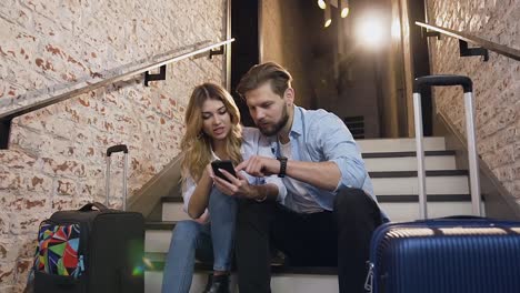 Angenehm-lächelnd-moderne-Mann-und-Frau-sitzen-auf-Treppen-in-Modehotel-mit-dekorierten-Ziegelwand-und-mit-Telefon