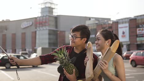 Divertido-joven-par-vietnamita-de-hombre-y-mujer-haciendo-selfie-con-peanapple-y-pan-largo-cerca-del-supermercado